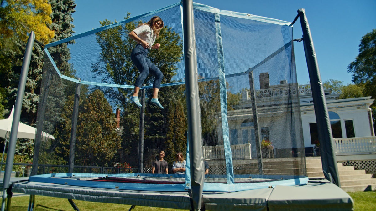Person jumping on a trampoline in a backyard with trees and a house in the background