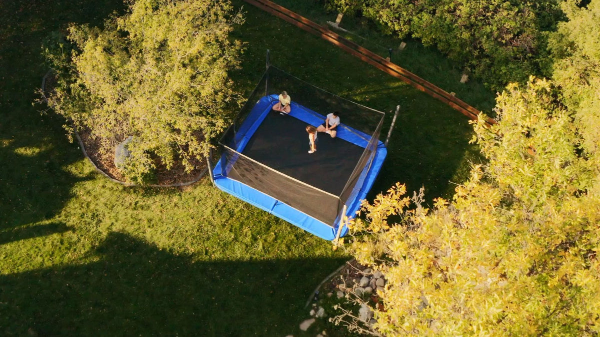 Two people on a blue inflatable trampoline in a grassy area with trees.