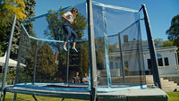 Person jumping on a trampoline in a backyard with trees and a house in the background