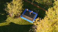 Two people on a blue inflatable trampoline in a grassy area with trees.